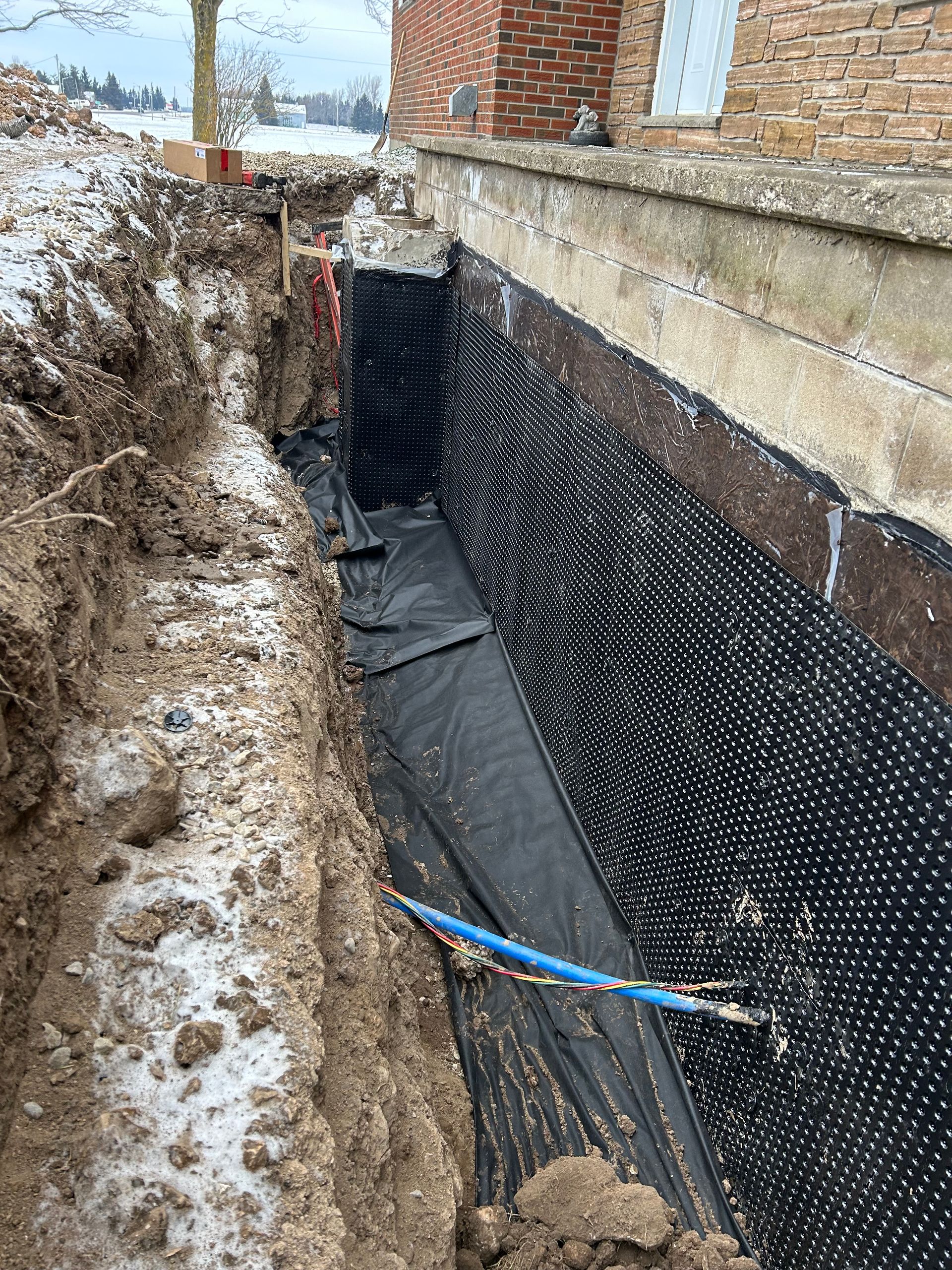 Exterior view of a foundation repair. Black dimpled drainage membrane and protective fabric installed along the foundation wall. Trench with snow.