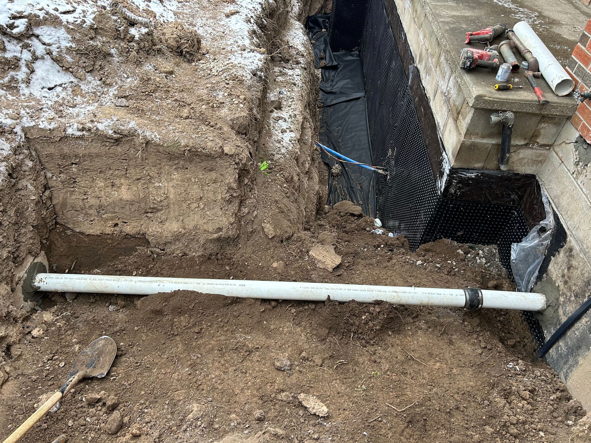 Construction trench with white pipe, soil, black barrier, near brick foundation. Snow present.
