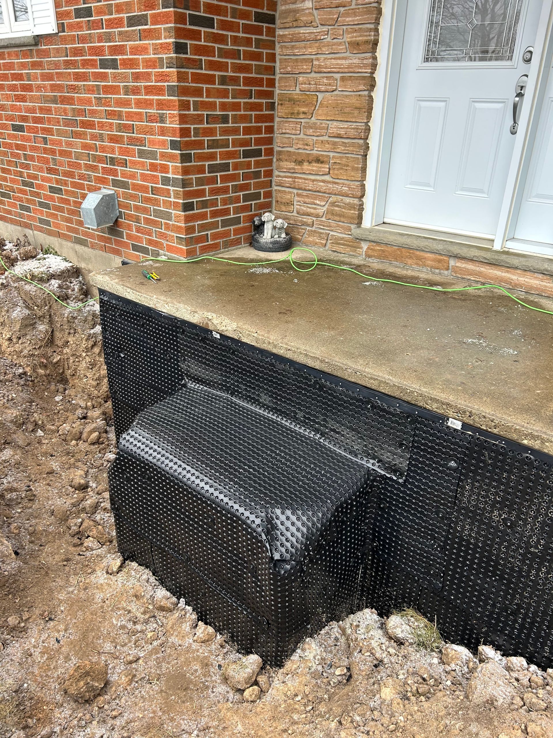 Foundation wrapped in black drainage membrane next to a brick wall and a concrete porch with a front door.