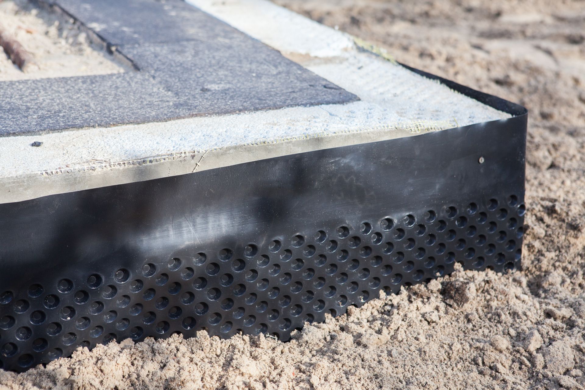 Black perforated drainage membrane around a concrete foundation, resting on sand.