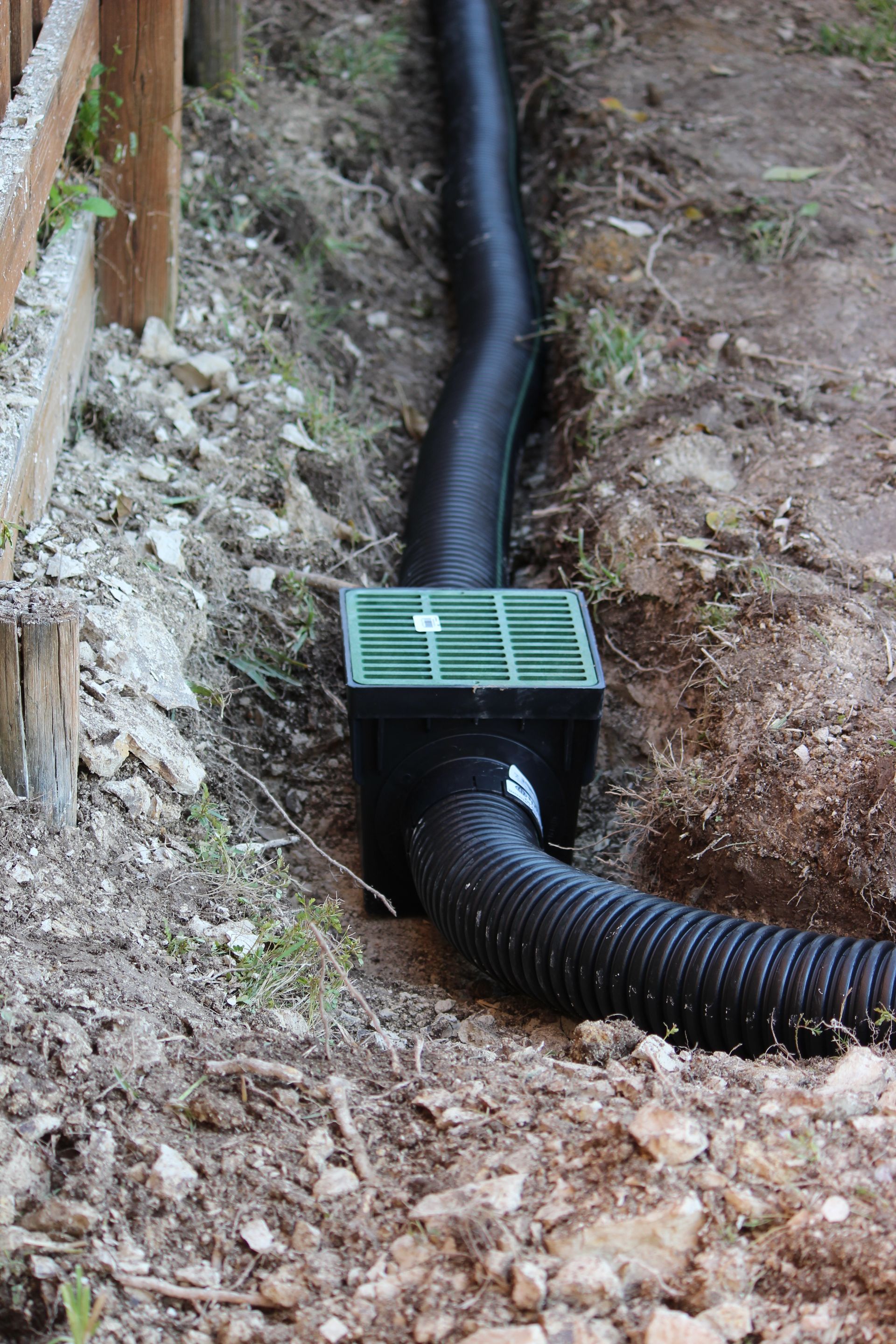 Black drainage pipe installed in a trench, connected to a green grate, next to a wooden fence.