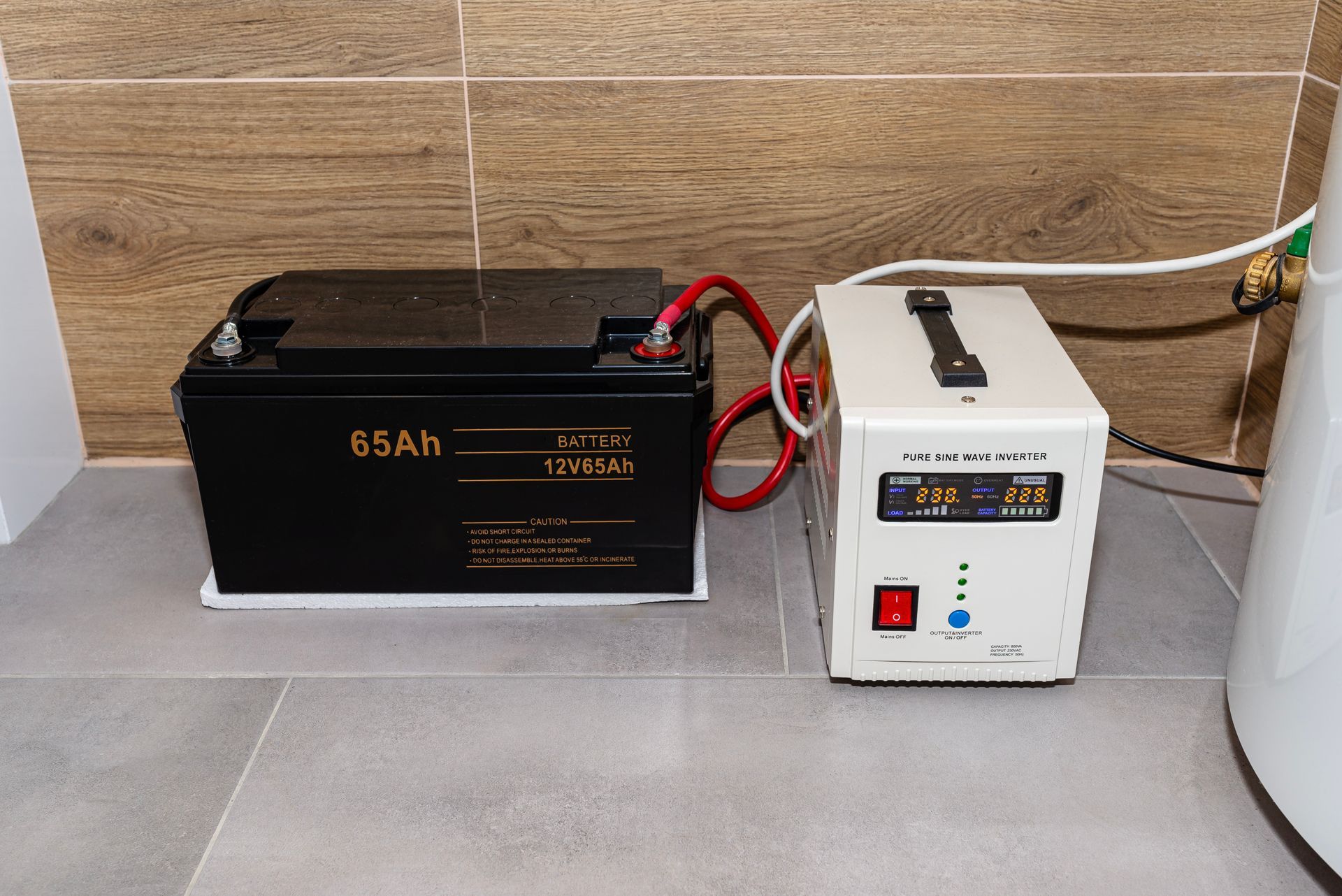 A battery connected to a white UPS device in a room with tile flooring and a wooden wall.