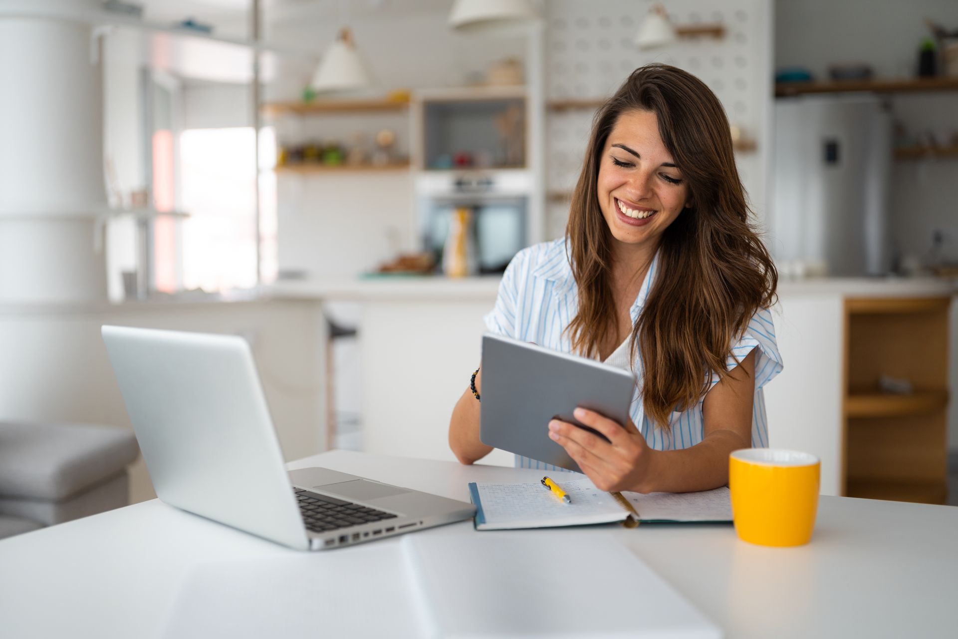 Woman smiles, working at a table with laptop, tablet, notebook, and coffee cup in a bright kitchen.