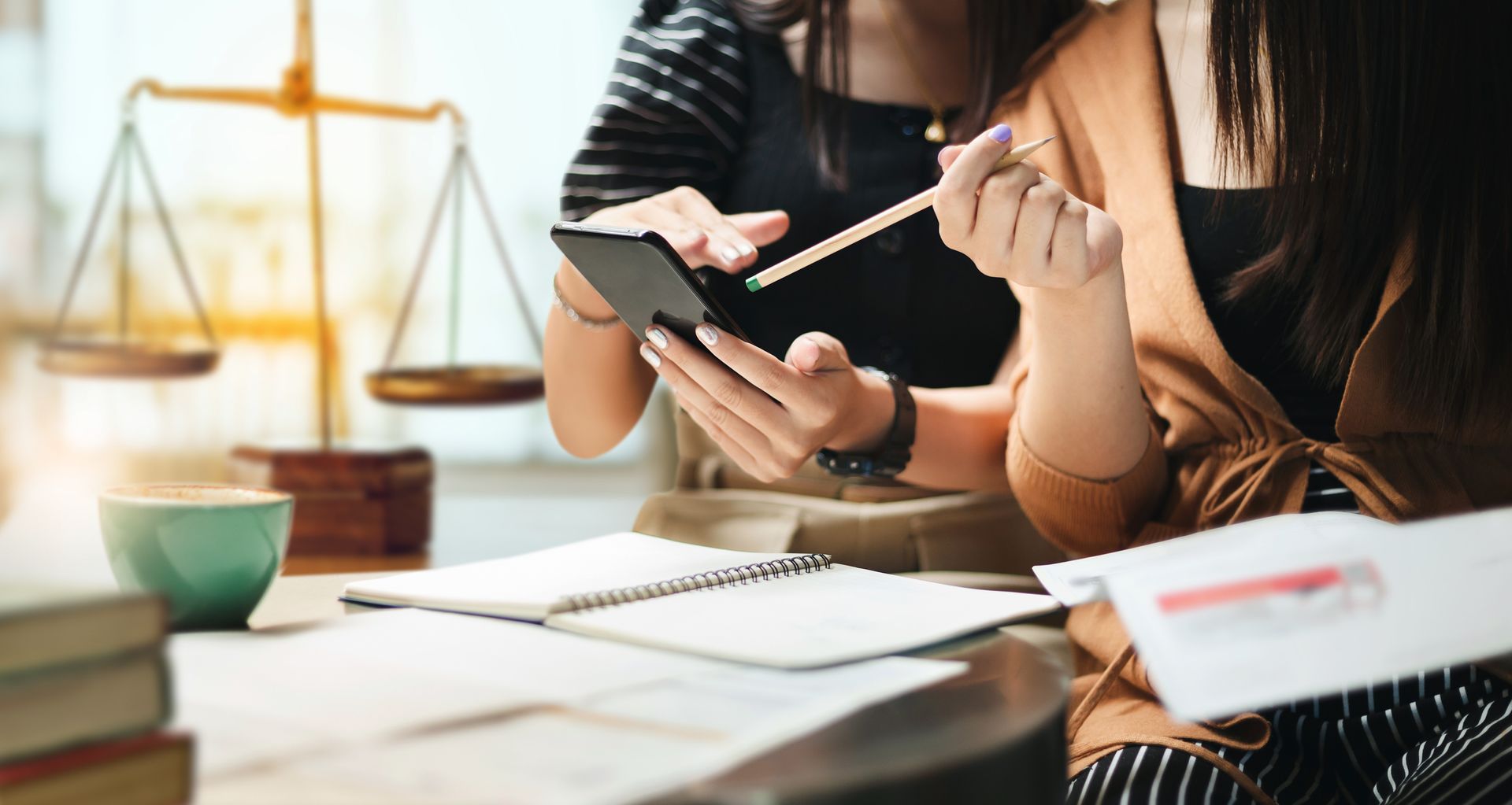 Two people at a desk review a phone, paperwork, and notebook, with a scale of justice in the background.