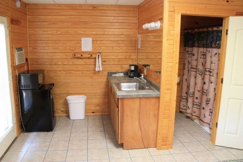 Small kitchen with wood-paneled walls, sink, mini fridge, trash can, and a shower stall in the corner.