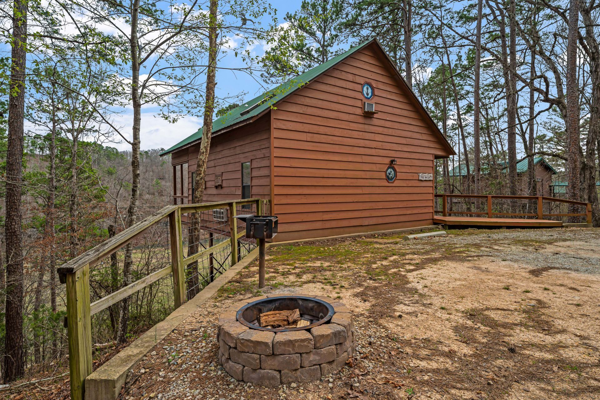 Wooden cabin on a wooded hillside with a fire pit and deck railing