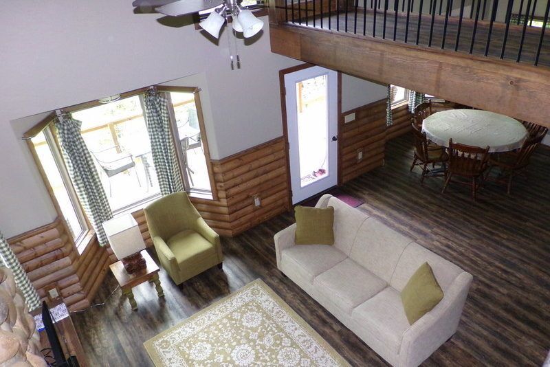 Cozy living room with beige sofa, green chairs, dining table, wood walls, and large windows