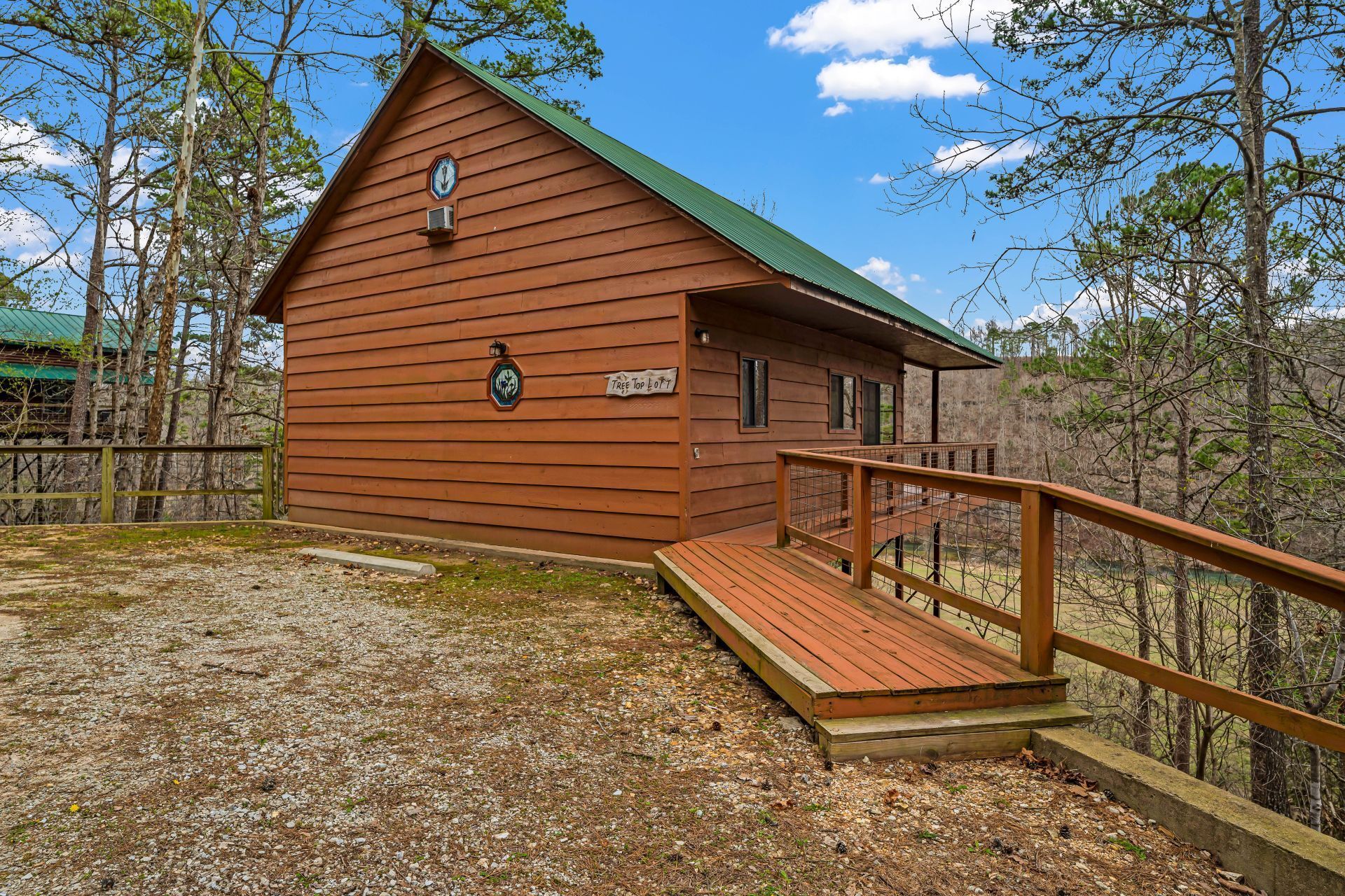 Brown cabin with green roof and wooden ramp on a wooded hillside under a blue sky