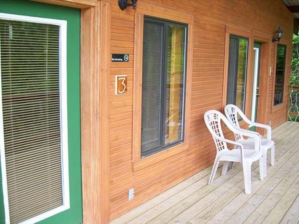 Wood-paneled porch with green-trimmed windows and two white plastic chairs on a wooden deck.