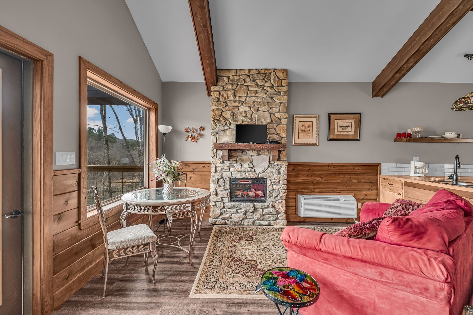 Cozy living room with stone fireplace, red sofa, round table by window, and wood ceiling beams