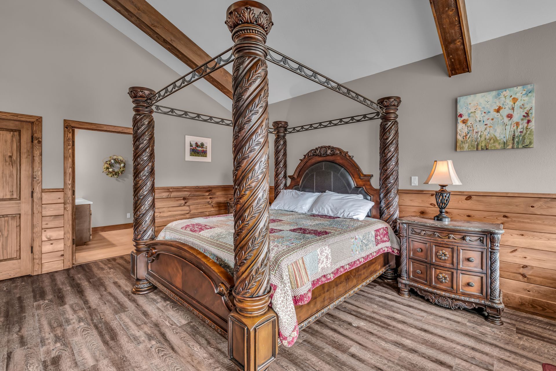 Rustic bedroom with a four-poster bed, wood beams, nightstand lamp, and wood-paneled walls.