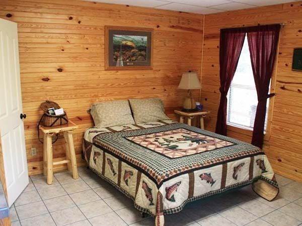 Cozy bedroom with wood-paneled walls, a bed, nightstand, lamp, and window with red curtains.