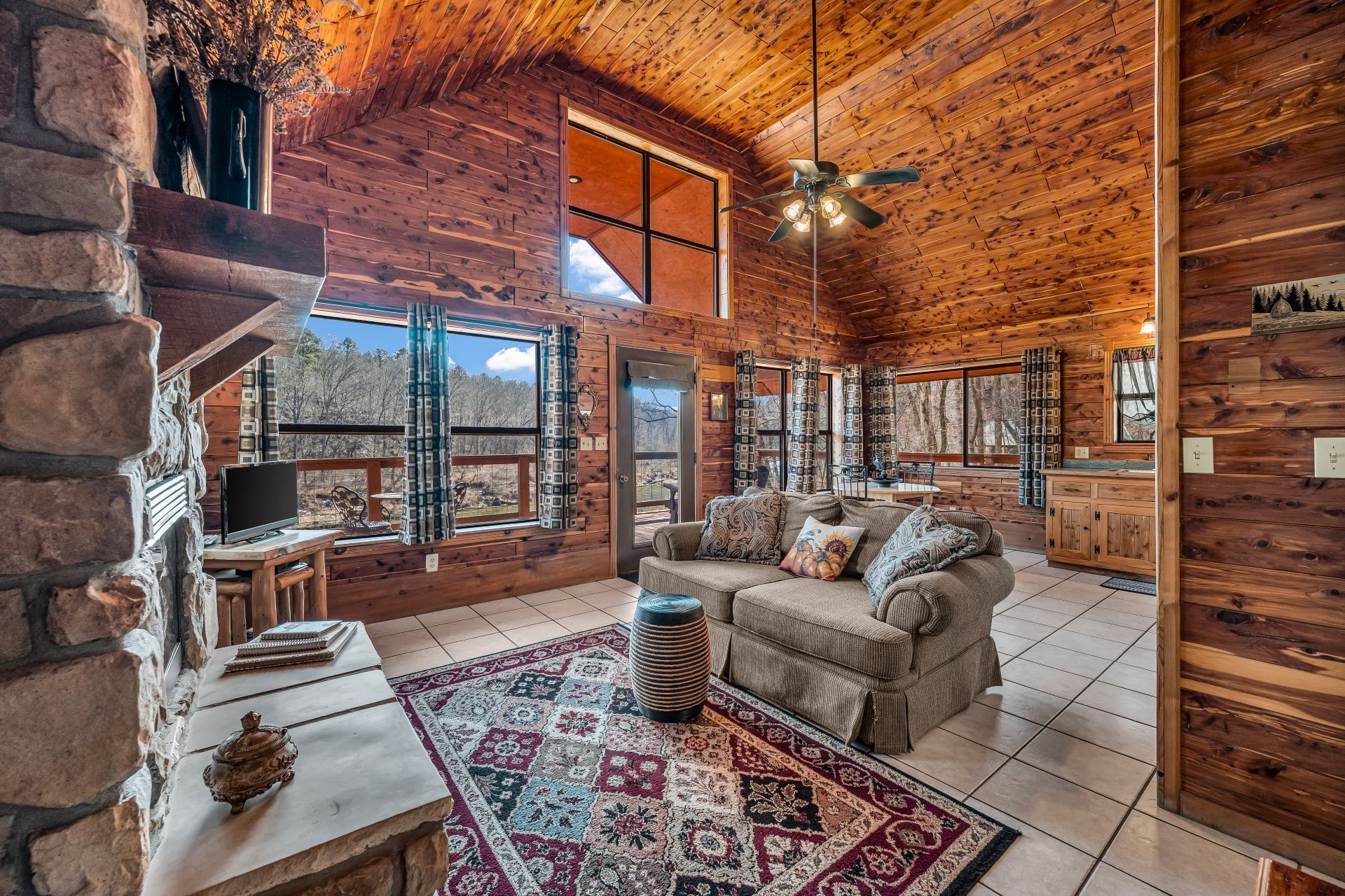 Rustic log cabin living room with vaulted wood ceiling, stone fireplace, large windows, and cozy sofa