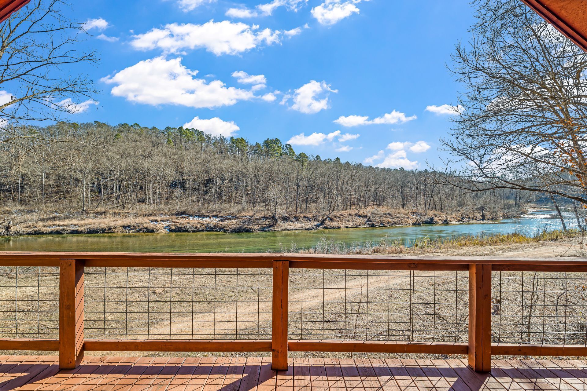 Wooden deck railing overlooking a calm river, leafless trees, and a blue sky with clouds.