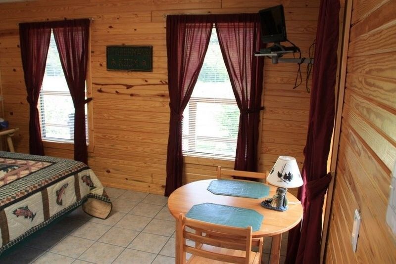 Cabin bedroom with wooden walls, bed, two curtained windows, and small round table with lamp