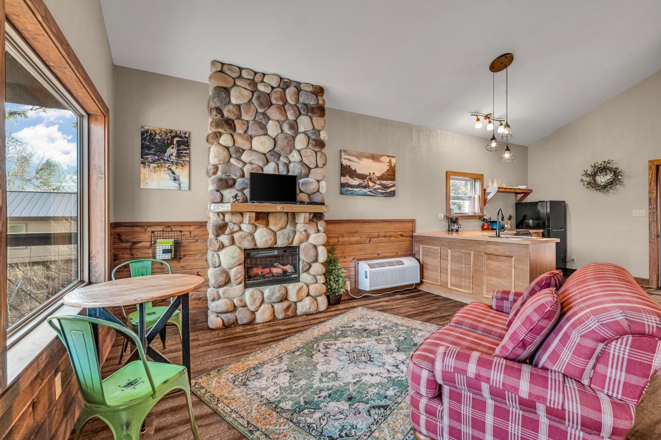 Cozy living room with stone fireplace, plaid sofa, colorful rug, and bright windows