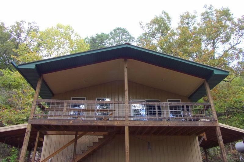 Wooden cabin deck with a metal railing under a green roof, surrounded by trees