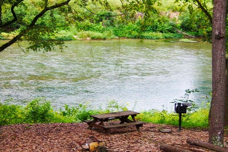 Wooded riverside with a picnic table and grill beside calm water
