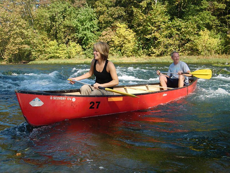 Two people canoeing in a red canoe on a river, with green trees along the bank.