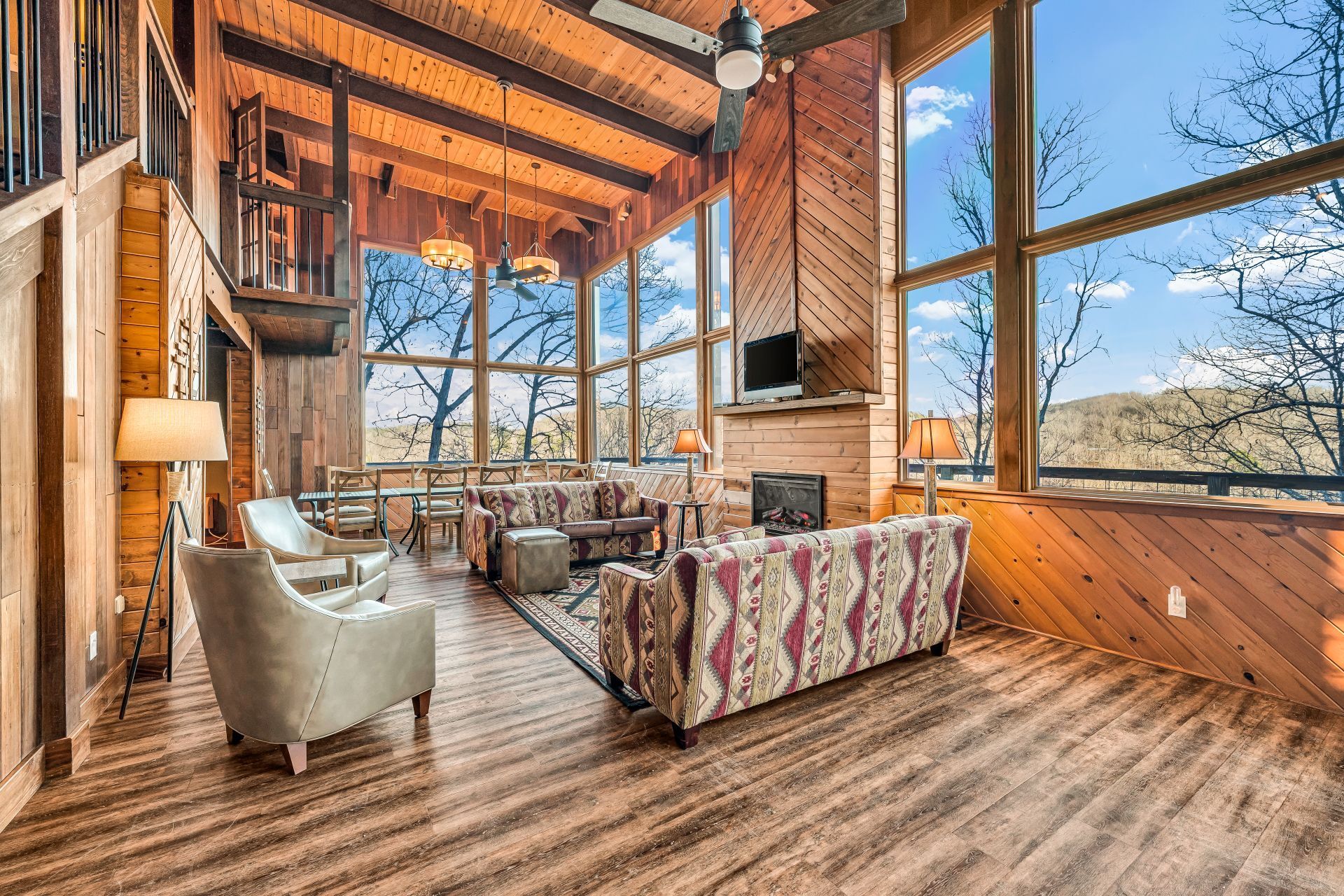 Rustic lodge living room with stone fireplace, large windows, and wood-beamed ceiling overlooking trees