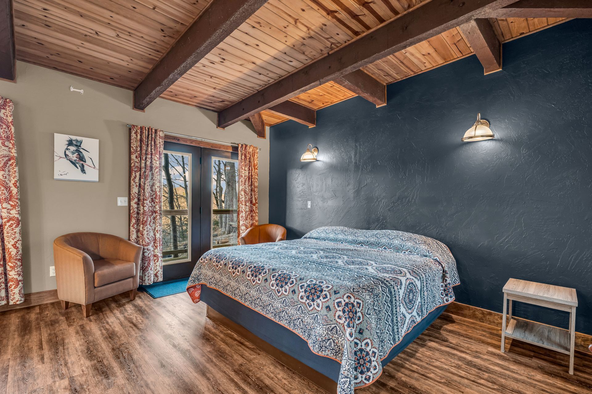 Rustic bedroom with wood-beamed ceiling, blue accent wall, patterned bedspread, and balcony doors