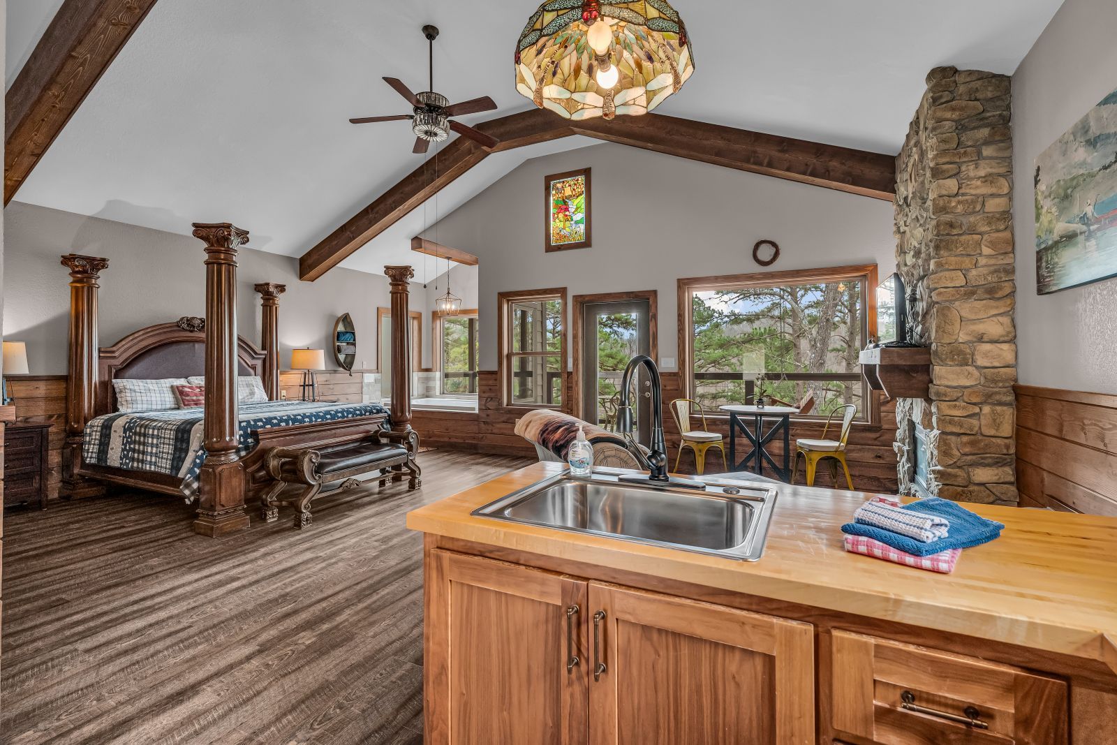 Open-plan rustic living room and kitchen with wood beams, stone fireplace, and large windows.