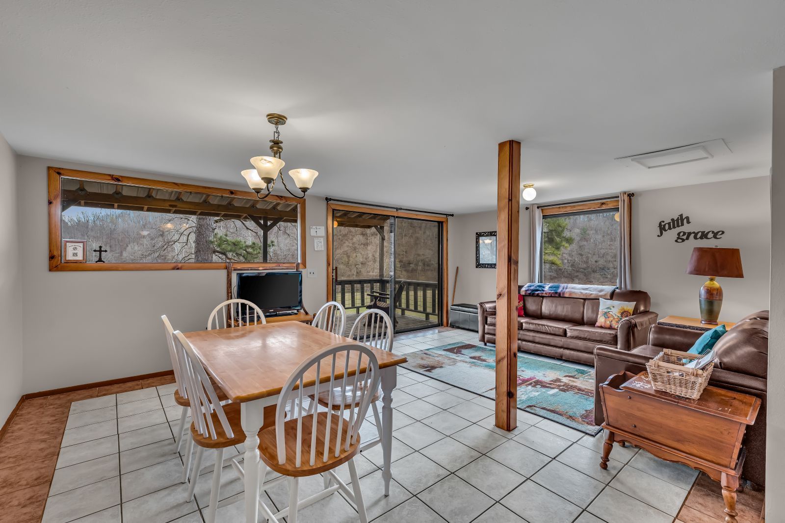 Bright dining and living room with tiled floor, wooden furniture, and large windows overlooking trees