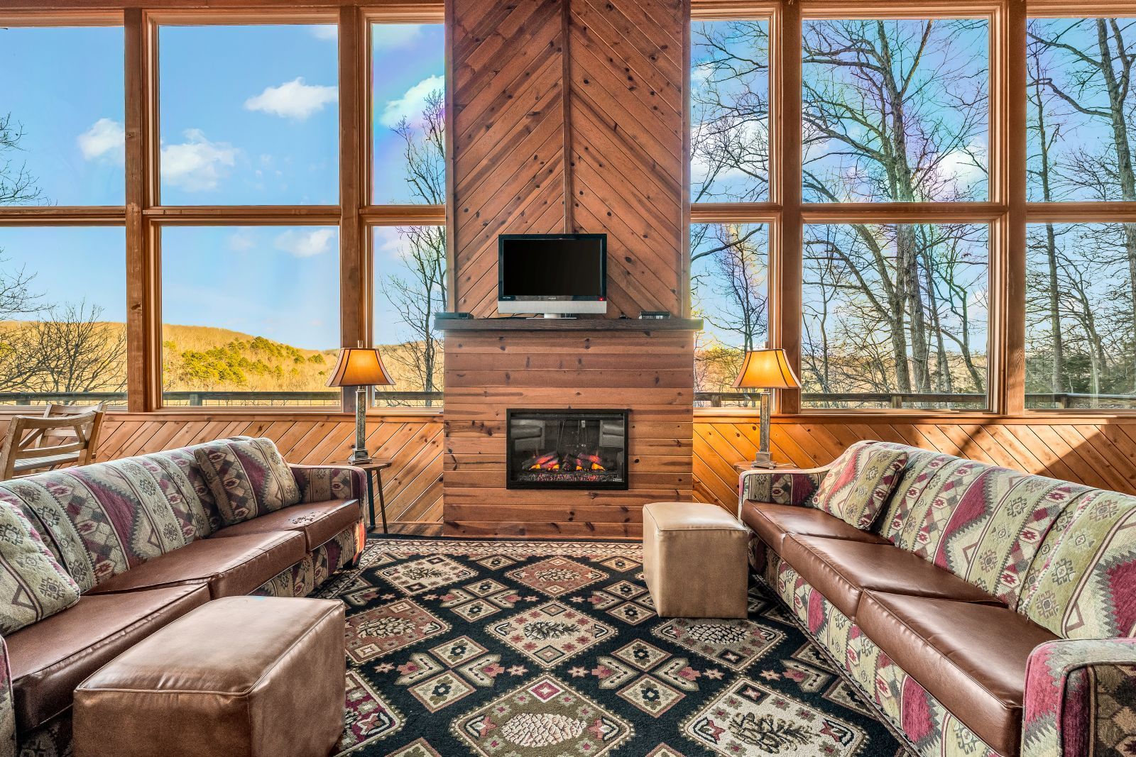 Cozy living room with leather sofas, patterned rug, fireplace, and large windows overlooking trees and sky