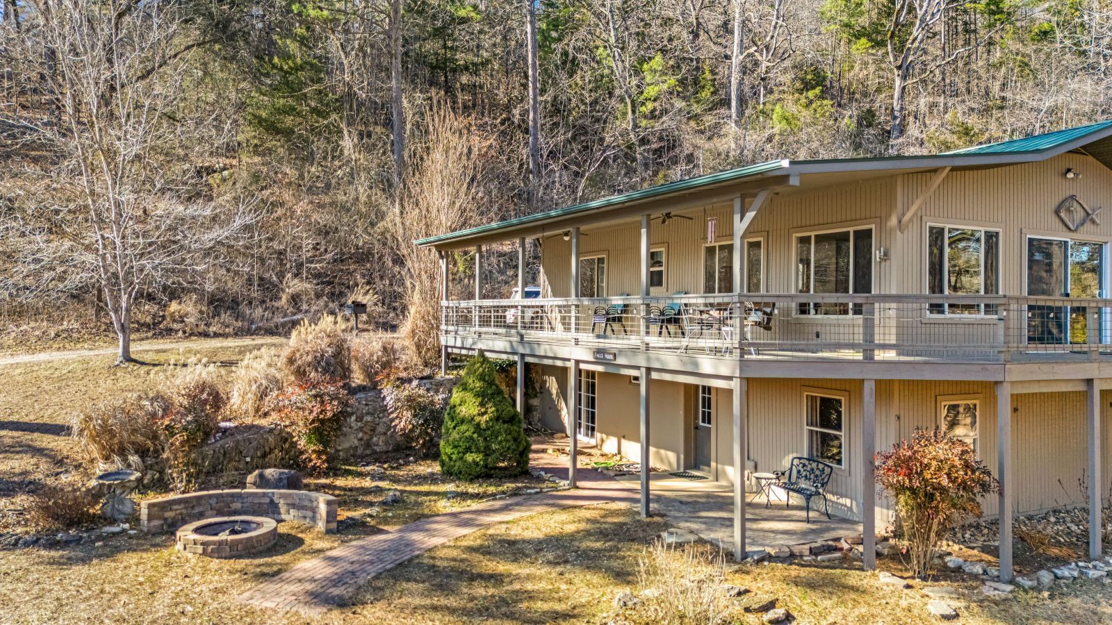A beige two-story house with a wraparound deck on a wooded hillside in autumn.