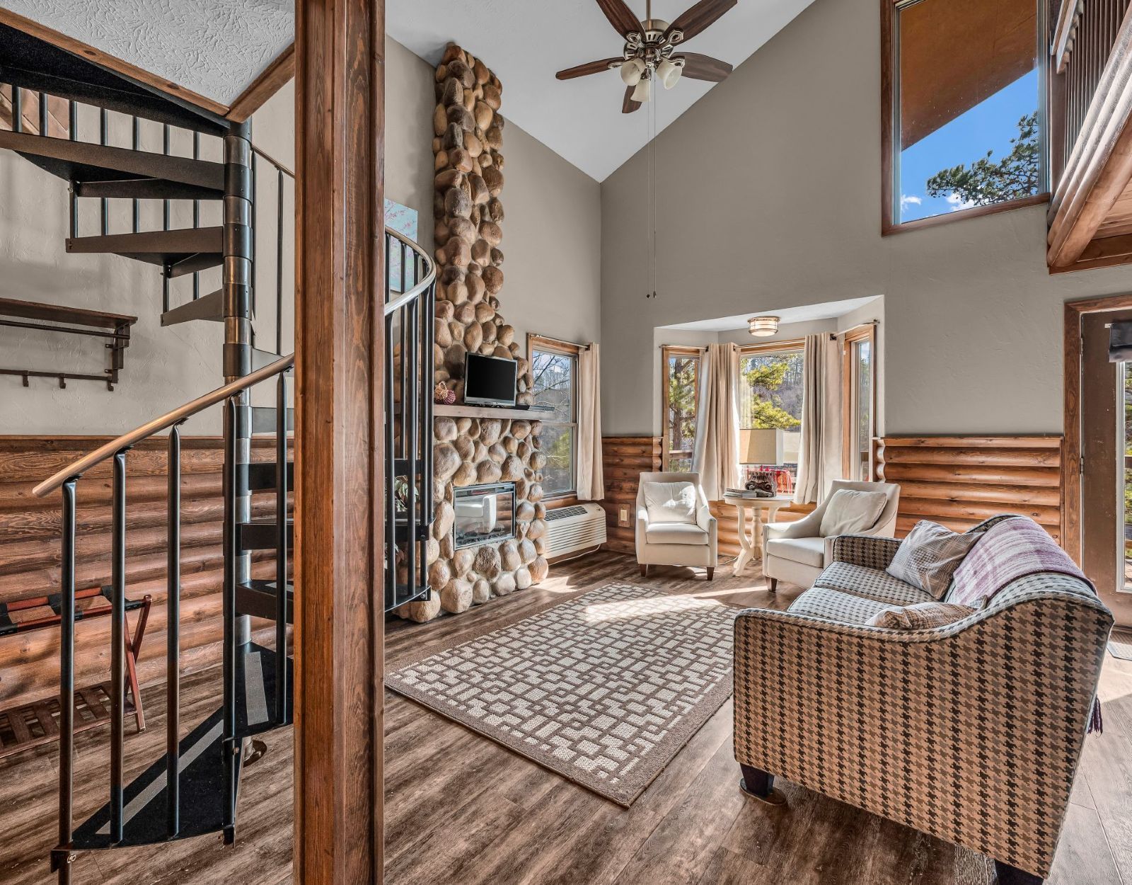 Cozy living room with wicker sofa, stone fireplace, staircase, and large windows