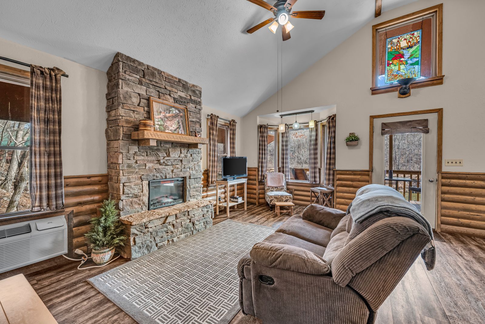 Cozy living room with stone fireplace, large windows, brown recliner, and ceiling fan.