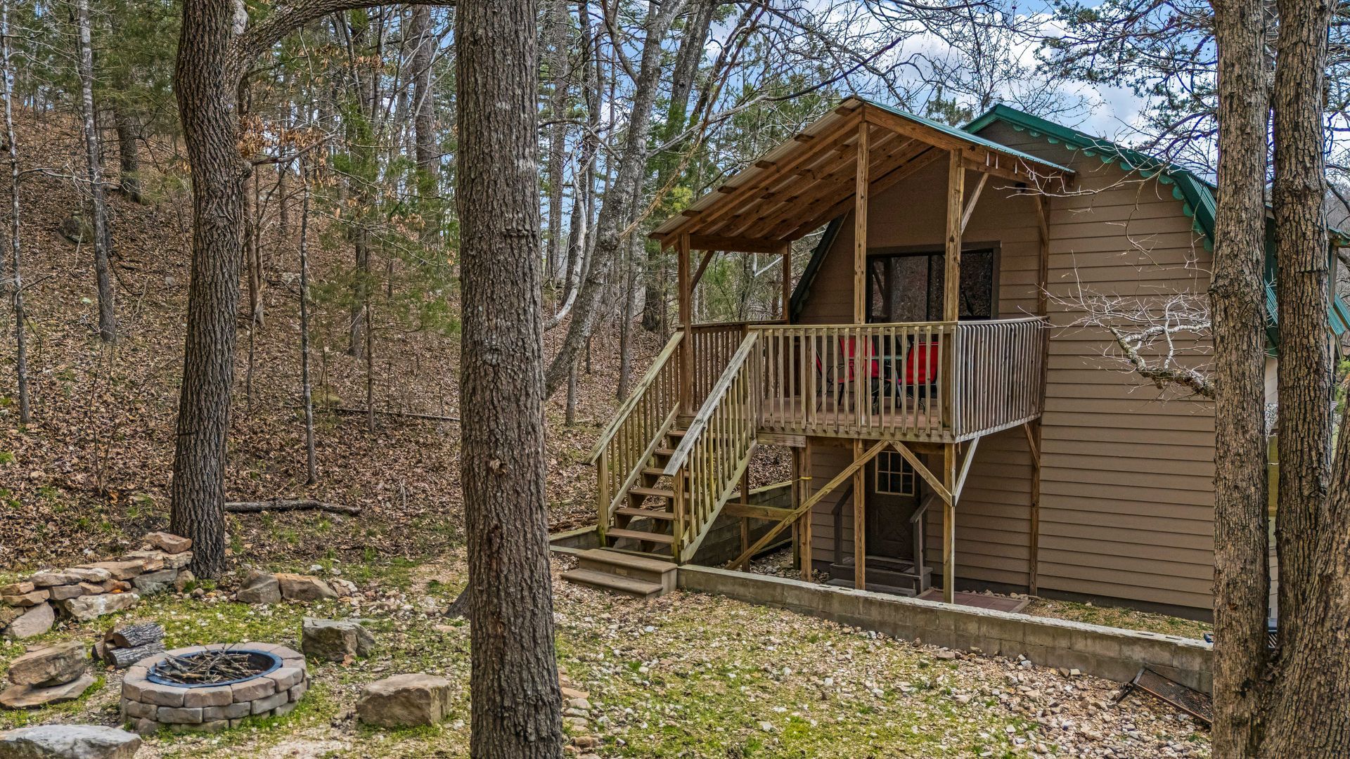 Small brown cabin with porch in a wooded area, with stairs and a tire fire pit nearby