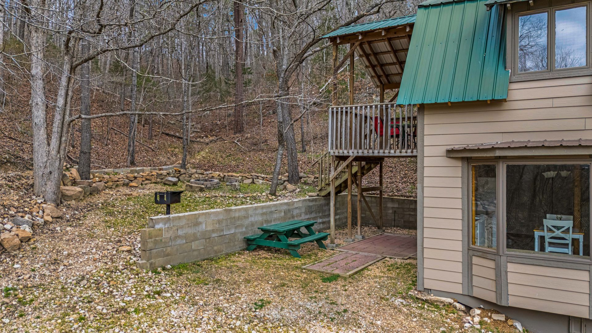Wooded backyard with a beige house, green awning, deck, and picnic table beside a stone retaining wall.