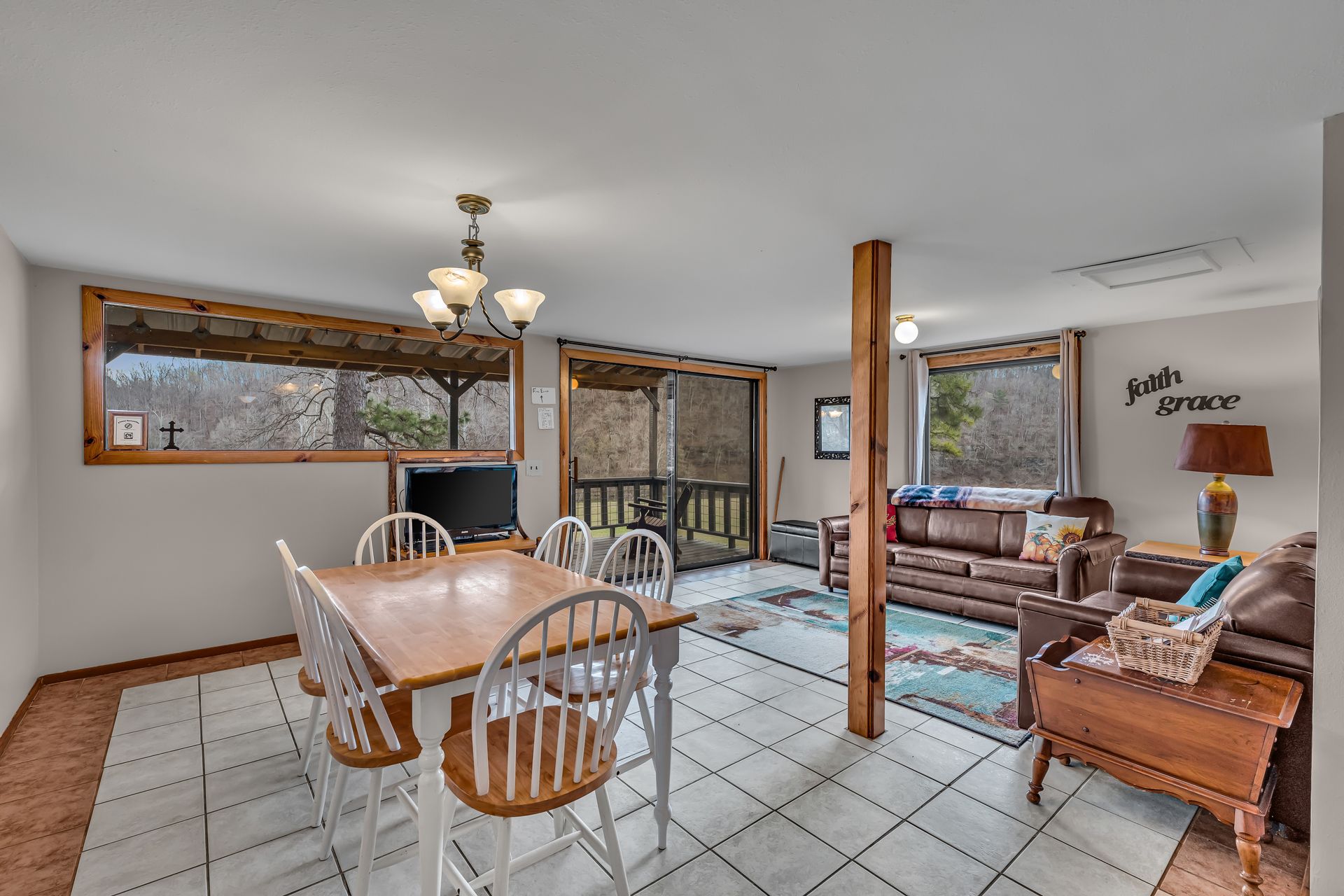 Open dining and living room with tiled floor, wooden table, chairs, and large windows overlooking trees.