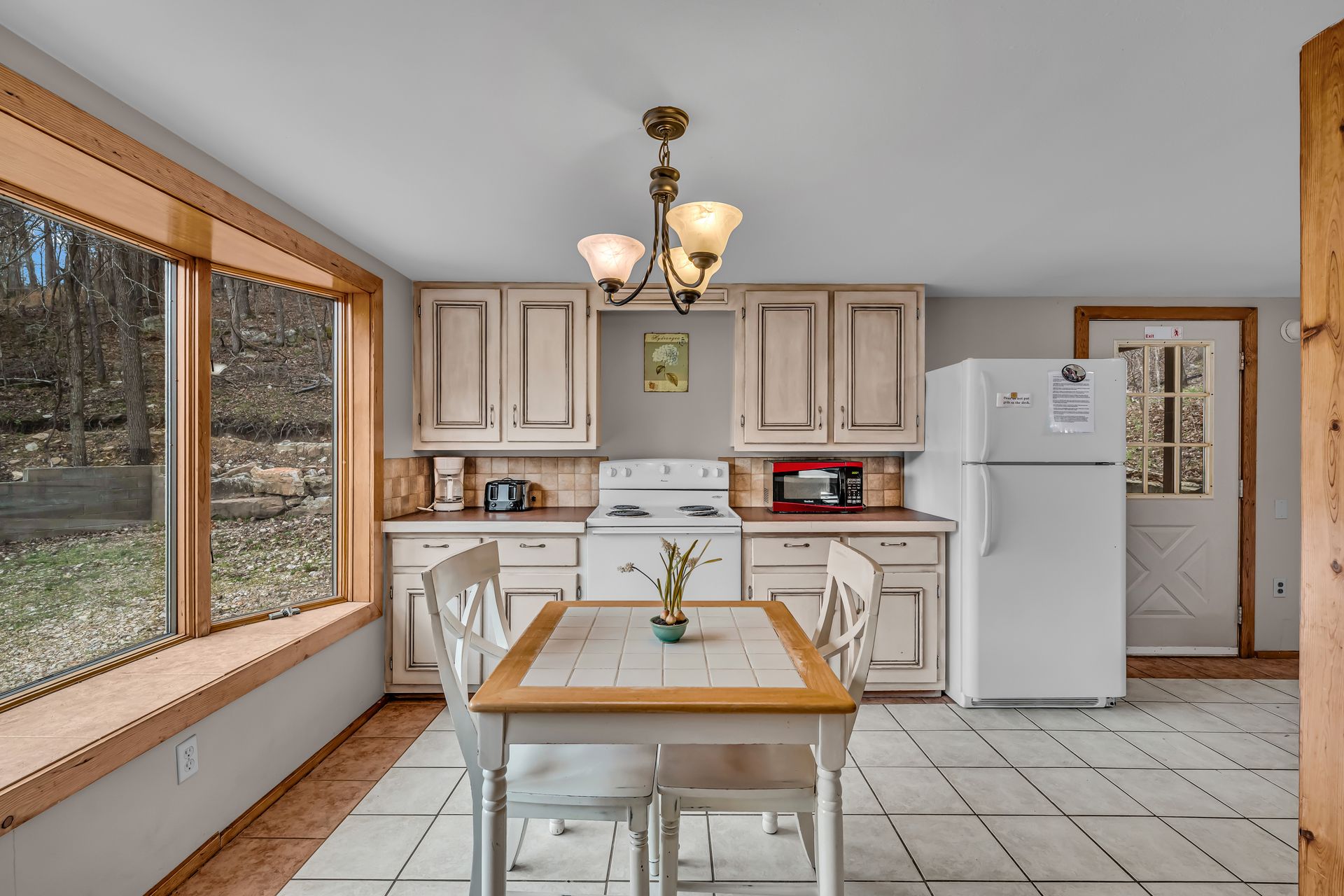 Bright kitchen with white cabinets, tiled floor, large window, dining table, and refrigerator
