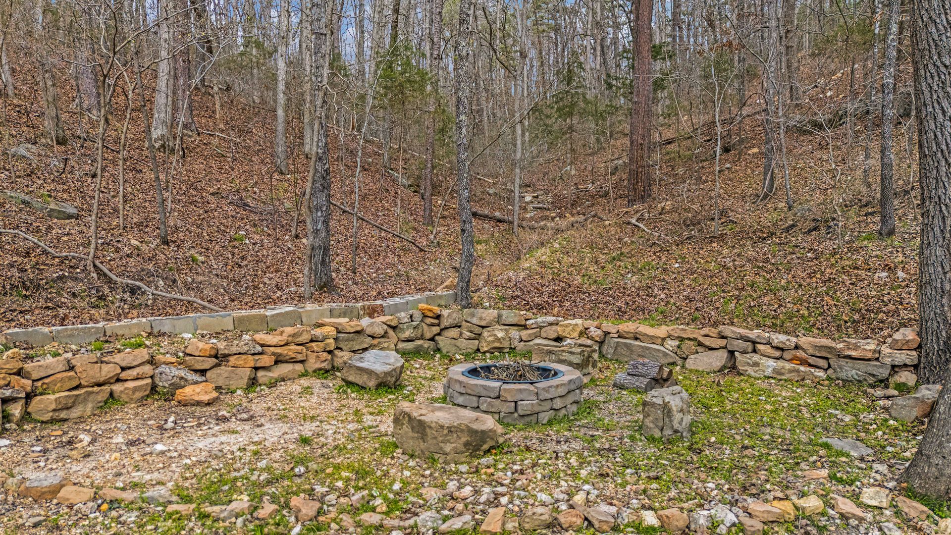 Stone fire pit in a wooded clearing with a low stone wall and bare trees in the background