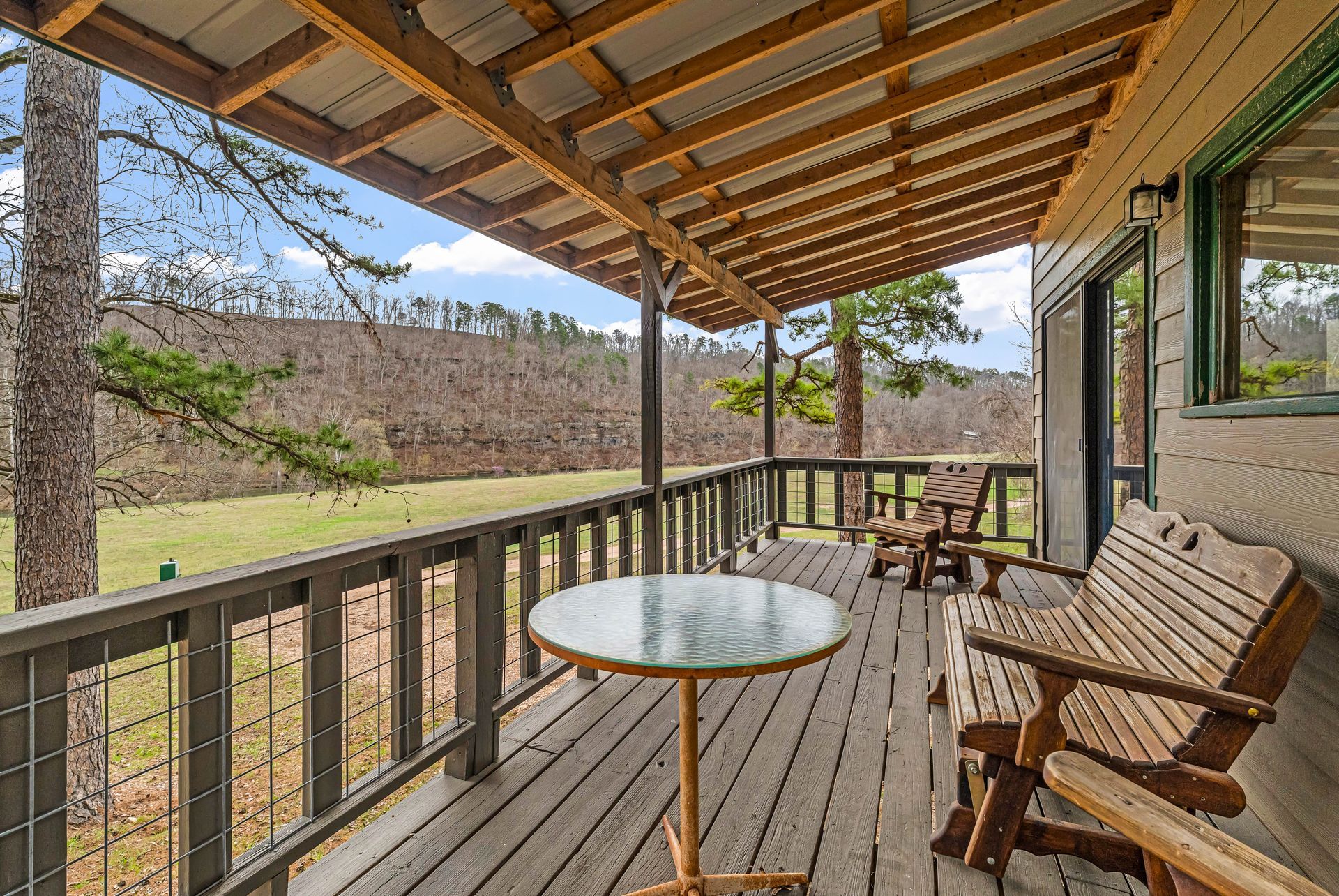 Wooden covered porch with railings, bench, and glass table overlooking a grassy yard and trees