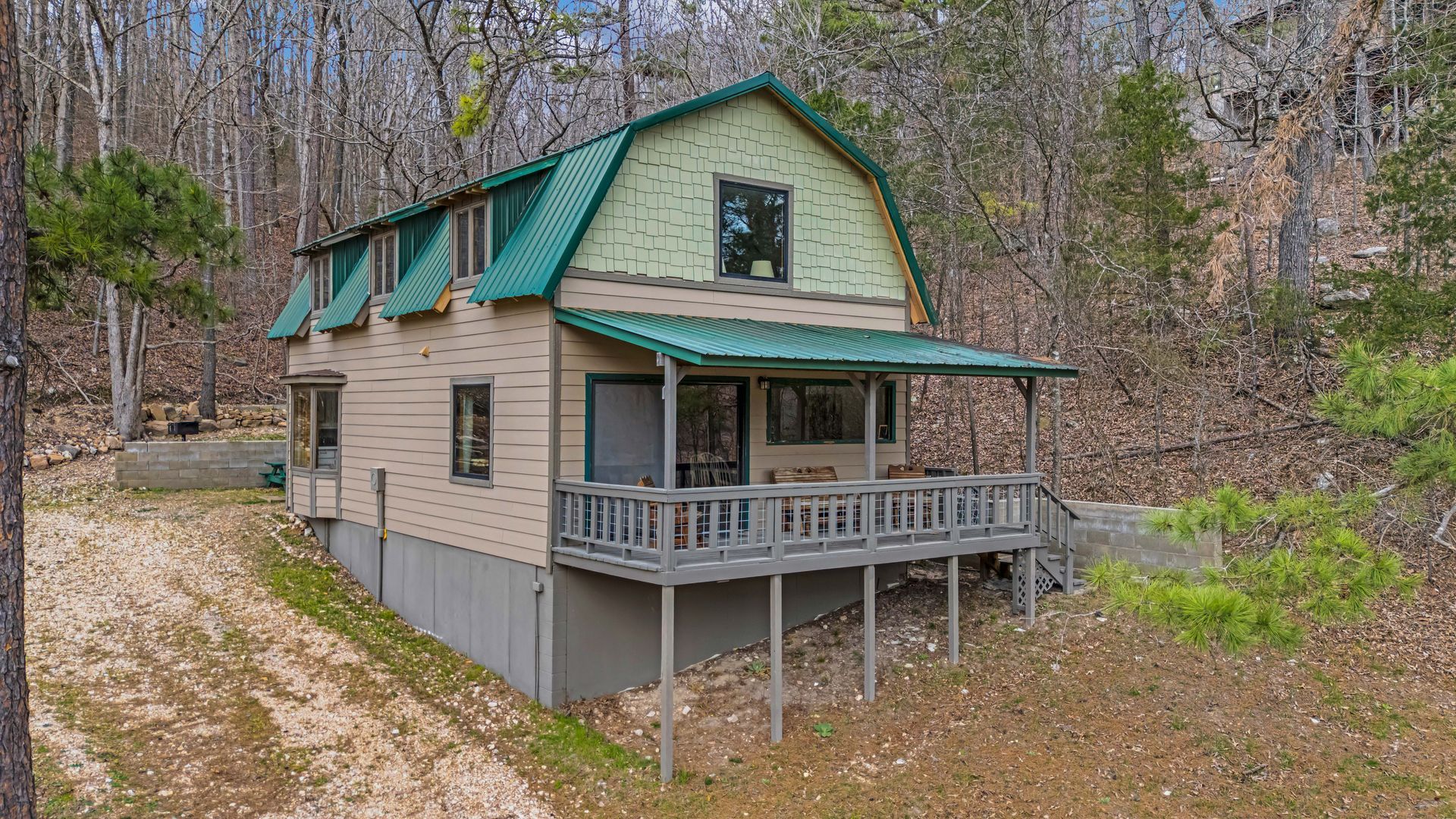 Small beige cabin with green roof and porch in a wooded hillside setting