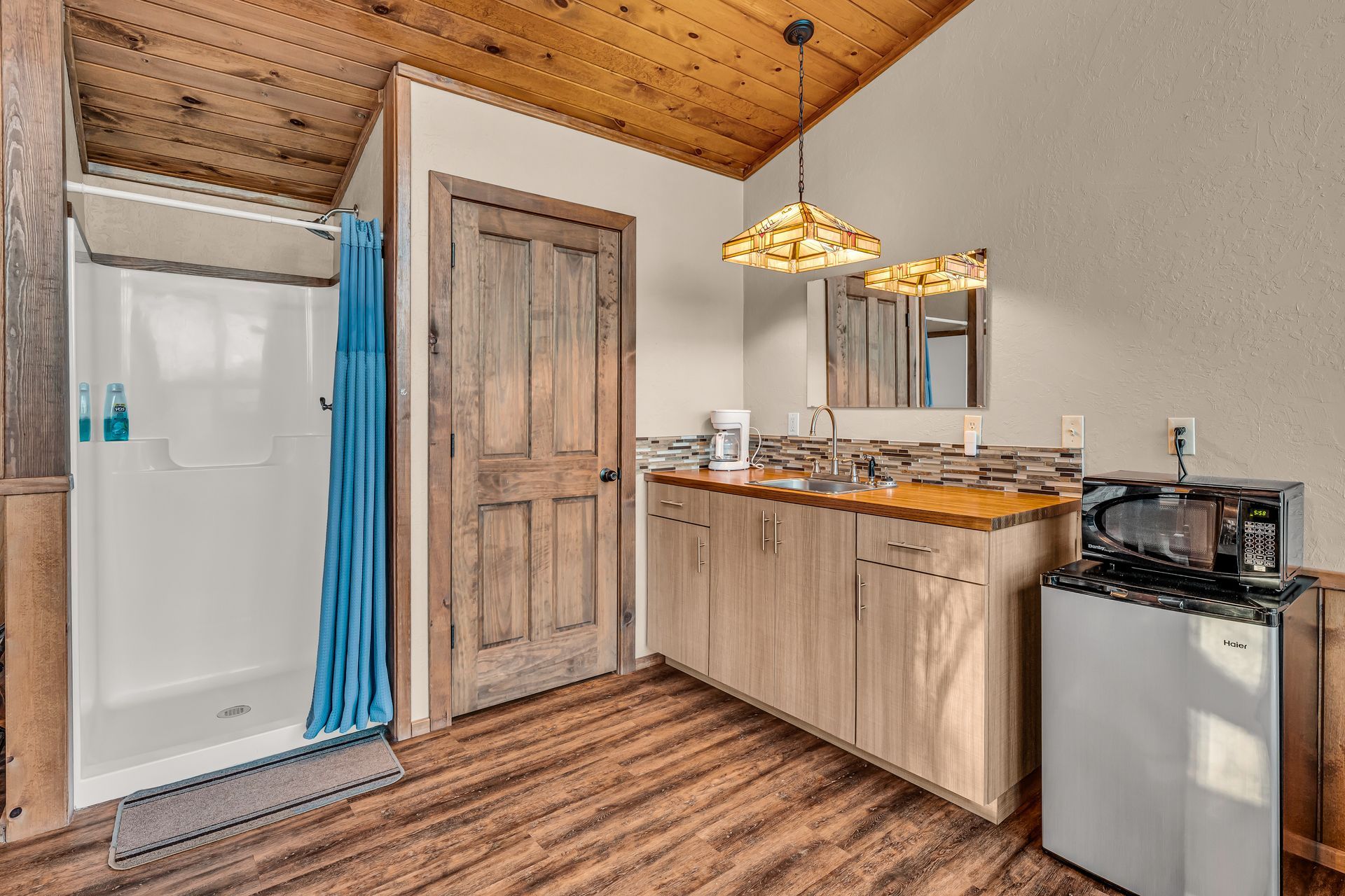 Cozy bathroom with shower, wood door, vanity, mirror, and pendant lights under a wood-paneled ceiling