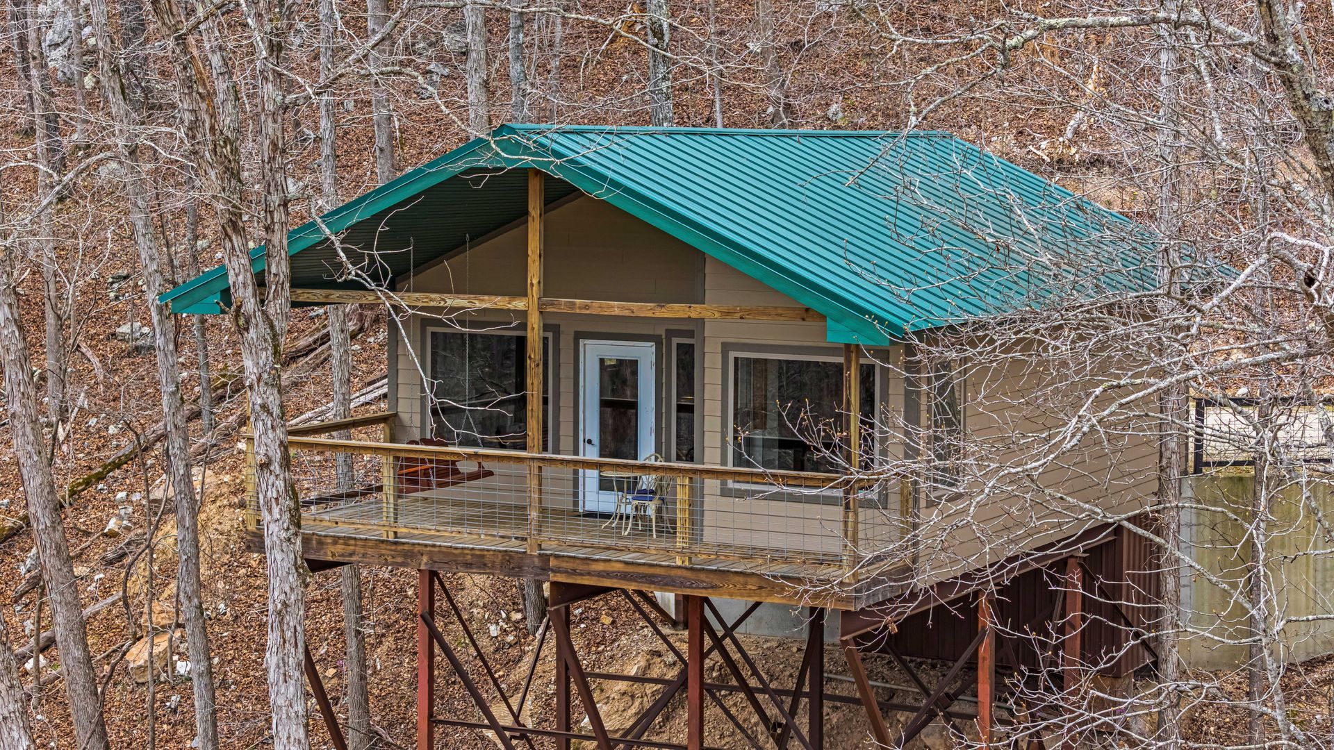 Small cabin on stilts with a teal roof in leafless woods, viewed from the side.