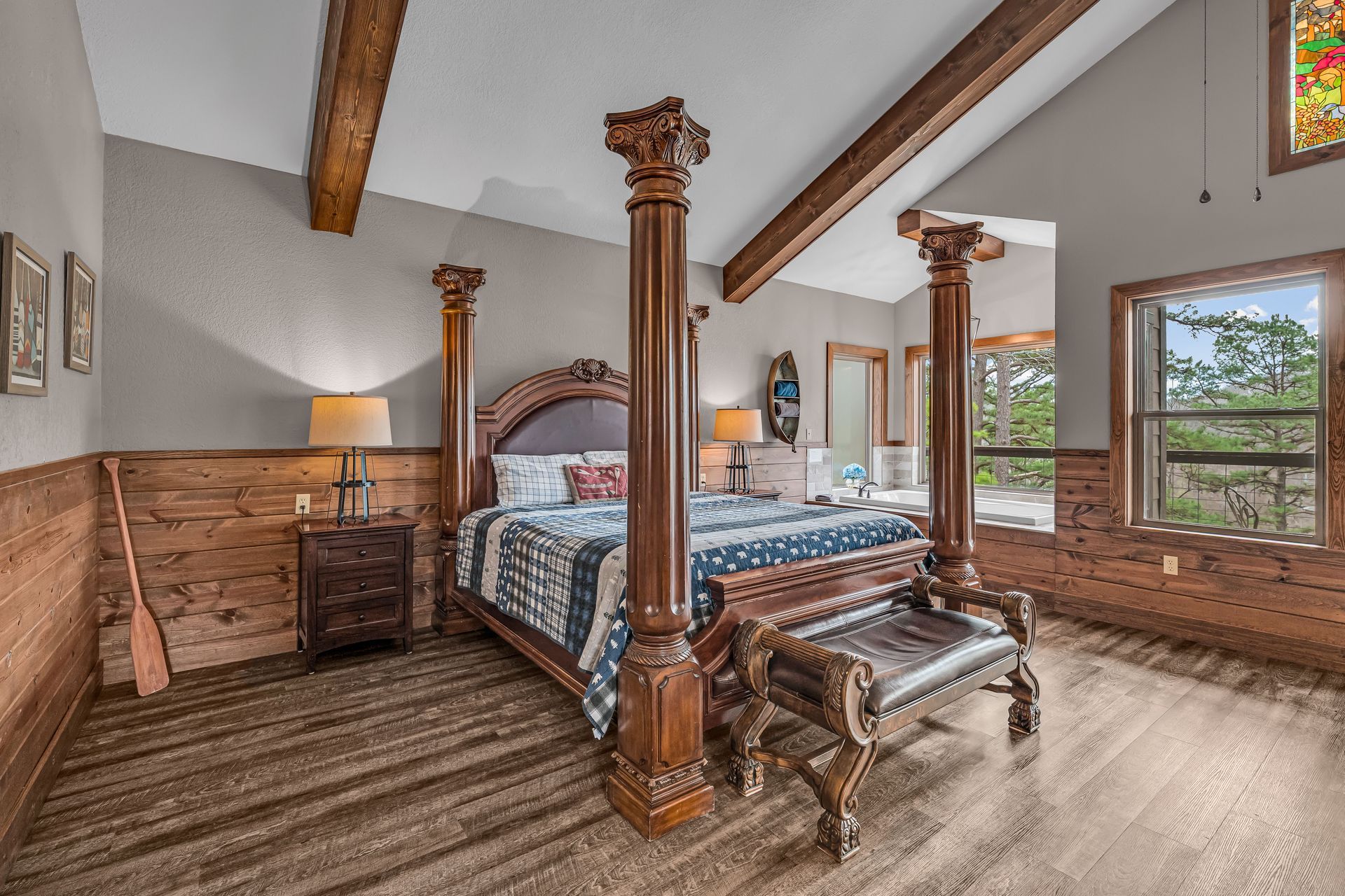 Rustic bedroom with four-poster bed, wood beams, and large windows overlooking trees.