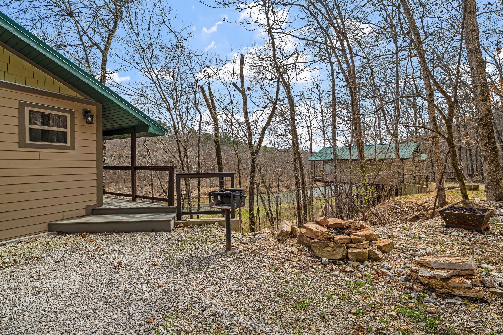 Gravel backyard beside a tan house, with a small deck, trees, and a stone fire pit.