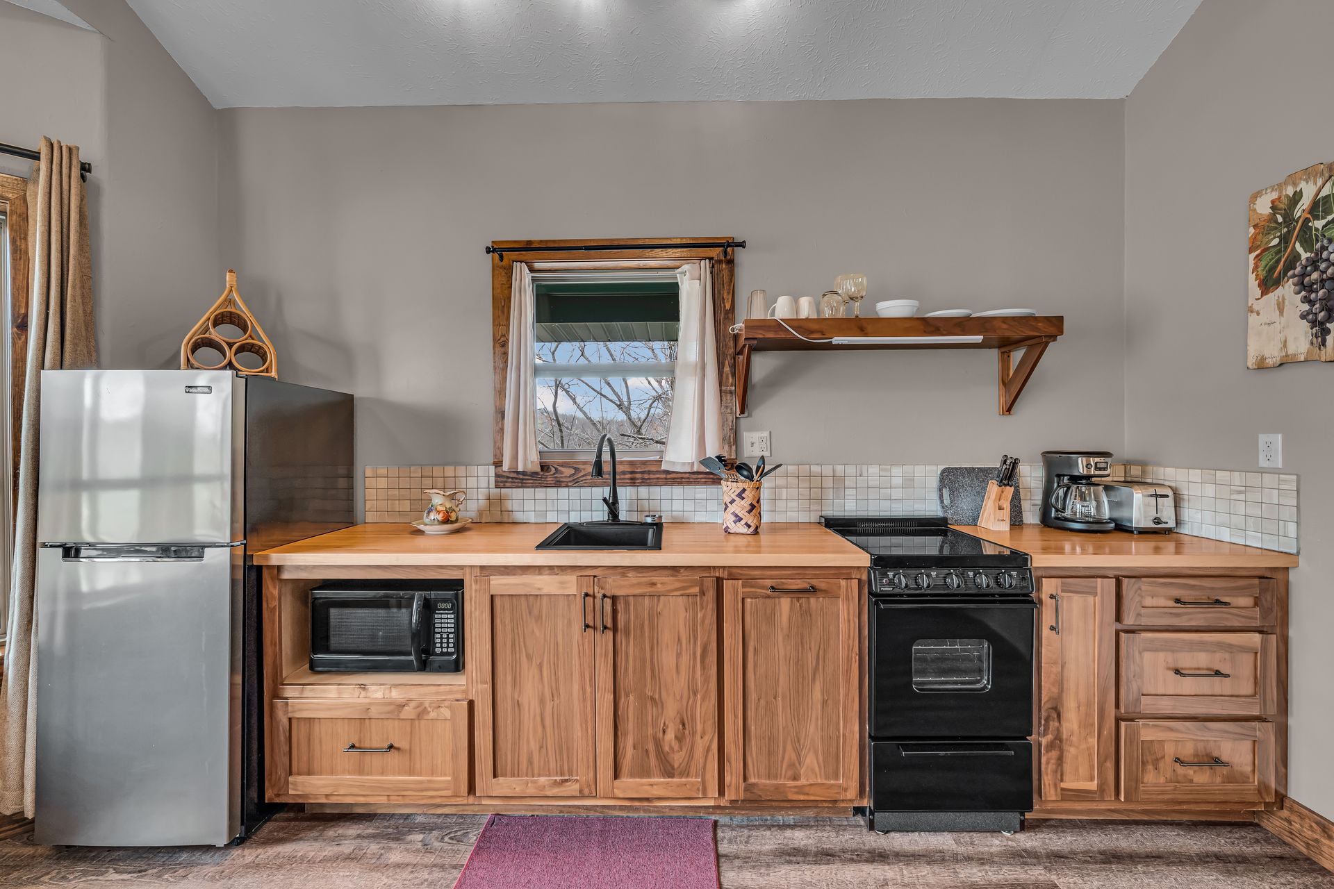 Small kitchen with stainless fridge, wood cabinets, sink, stove, and open shelf under a window.