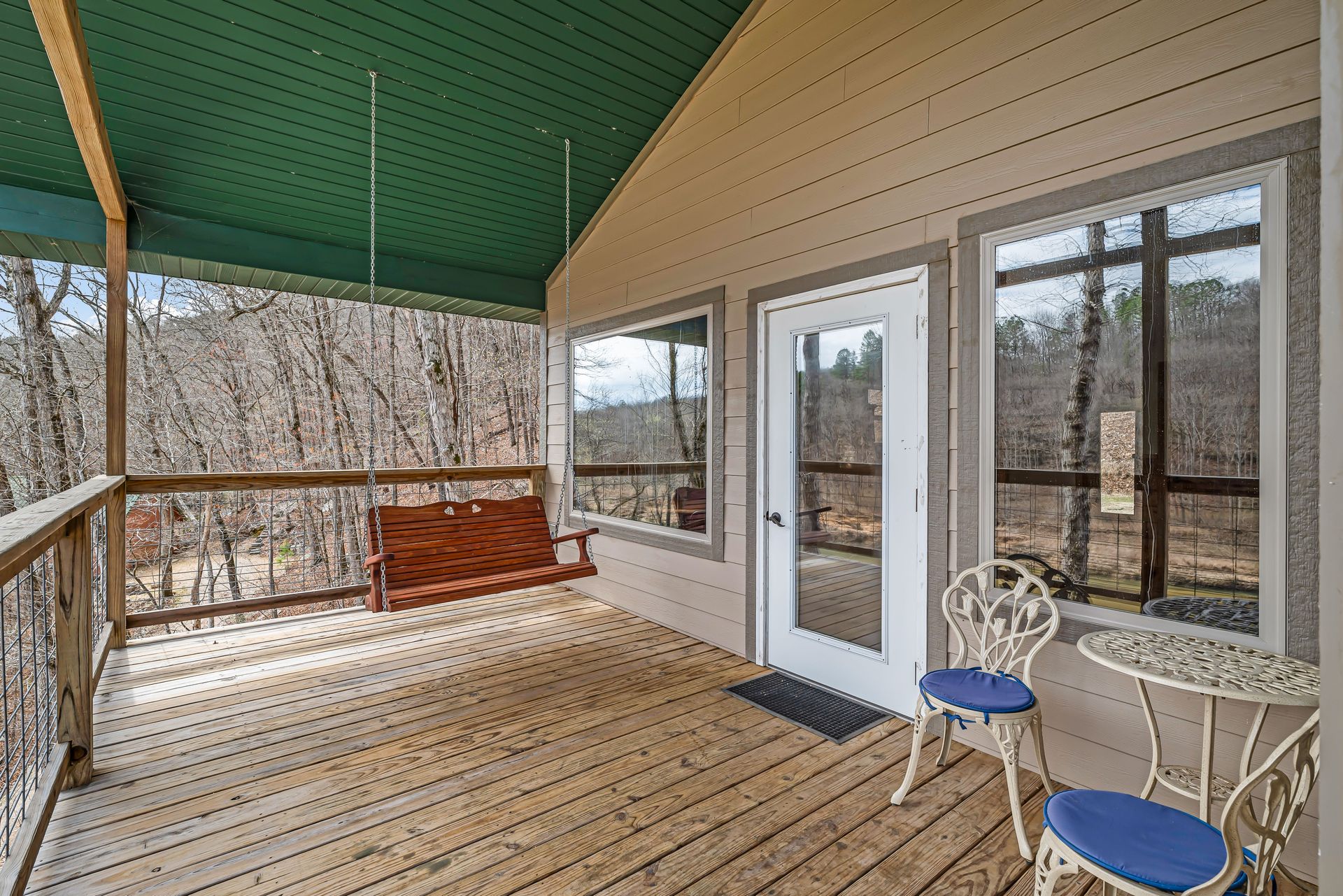 Covered wooden porch with a swing, white door, and two blue chairs overlooking leafless trees.