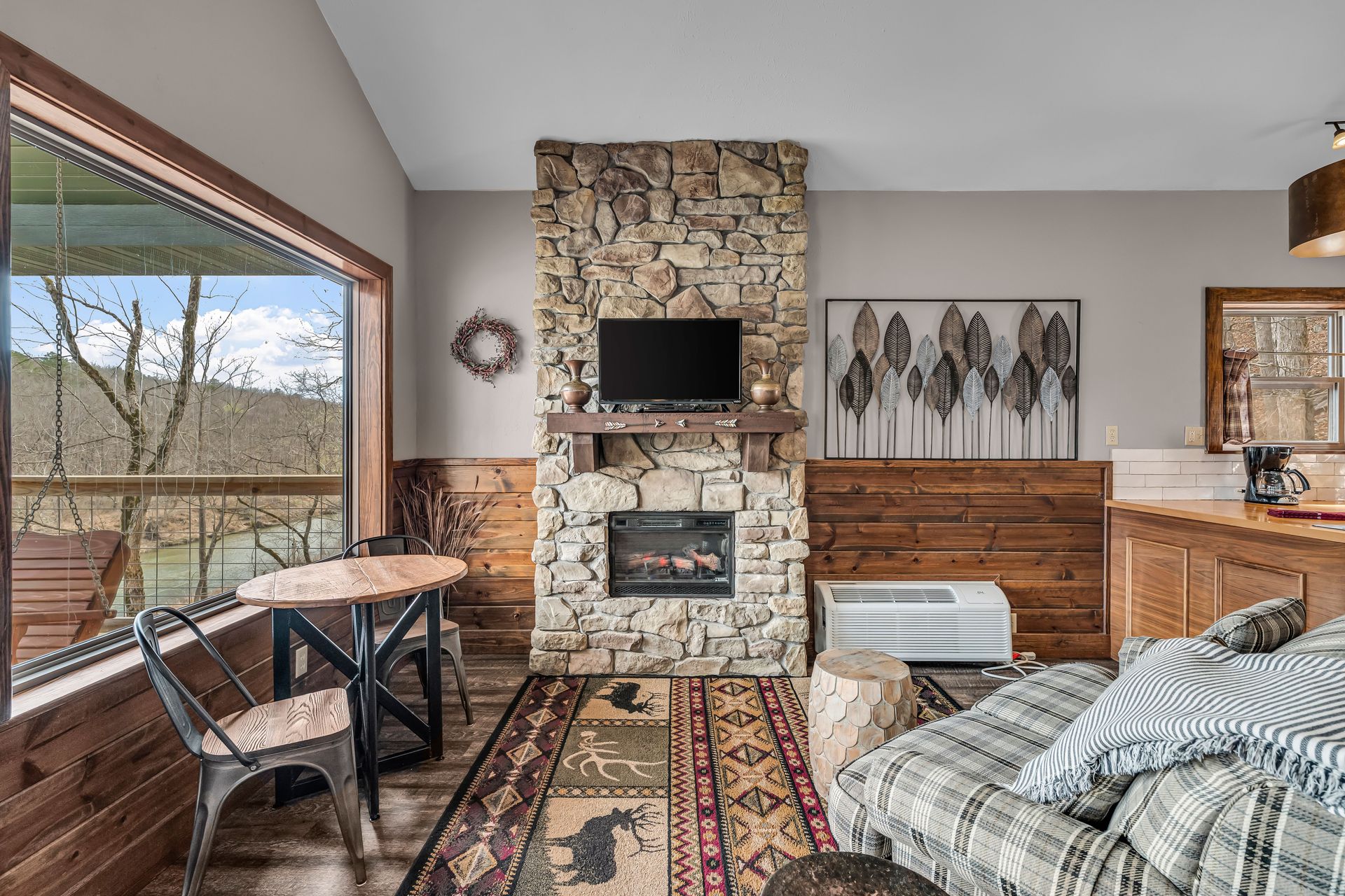 Cozy rustic living room with stone fireplace, wood paneling, patterned rug, and large window seating area