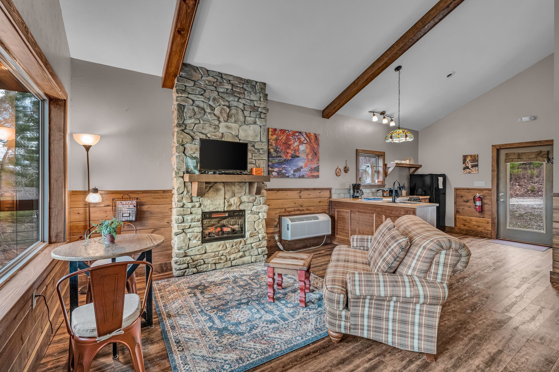 Spacious living room with stone fireplace, plaid sofa, wood beams, and dining area by large windows