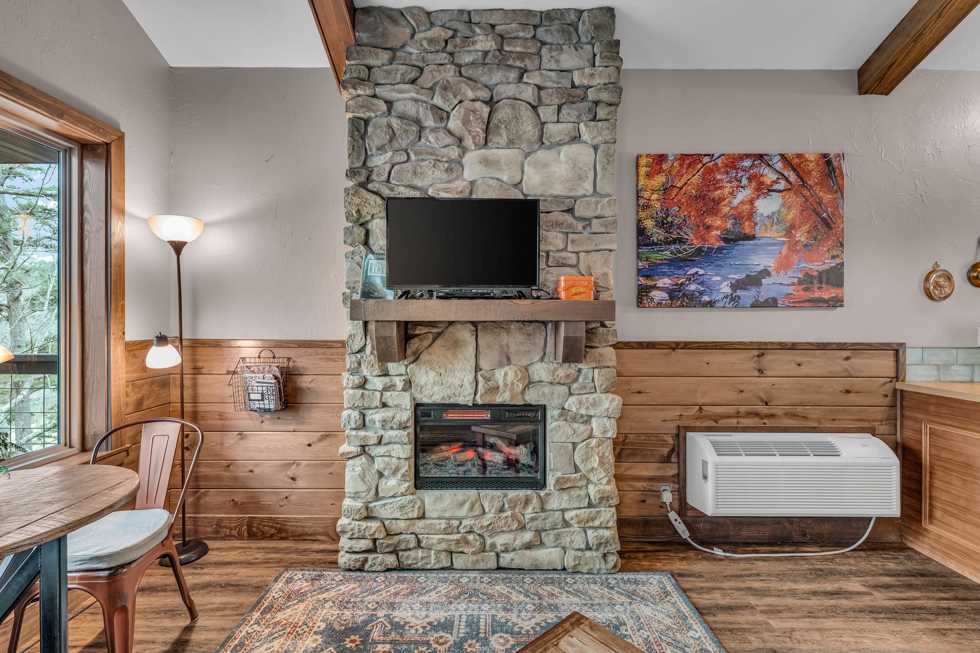 Cozy living room with stone fireplace, wood paneling, TV, colorful wall art, and a window seat