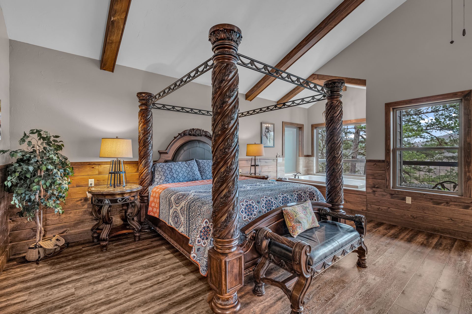 Spacious bedroom with a four-poster bed, wooden beams, a sitting chair, and large windows with forest views