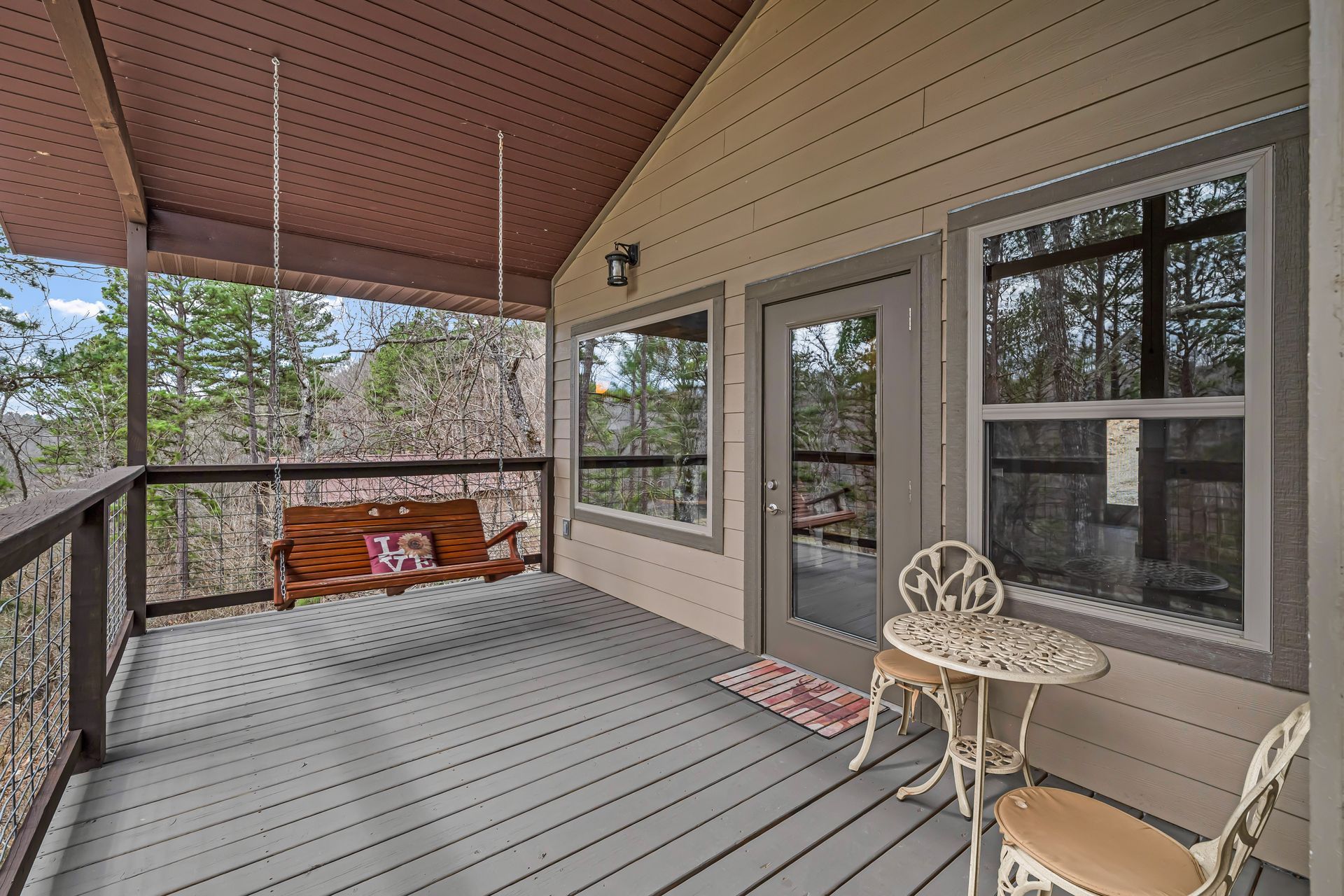 Covered porch with gray flooring, beige siding, windows, a glass door, and a small chair and table.