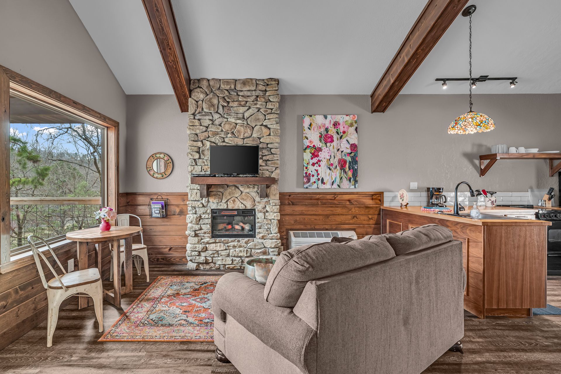Cozy living room with stone fireplace, wooden beams, sectional sofa, and kitchen open to the right.